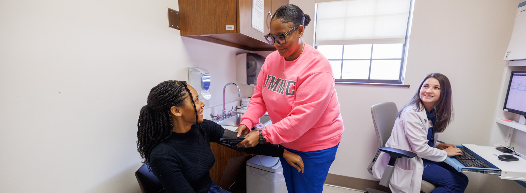 Provider taking patients blood pressure while another provider watches while sitting at a desk in front of a computer.